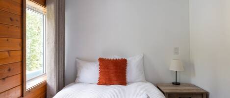 Ein Schlafzimmer mit einer Holzwand, einem Fenster, einem Bett mit weißer Bettwäsche und einem orangefarbenen Zierkissen, einem Nachttisch mit einer Lampe und einer Nachttischschublade.
