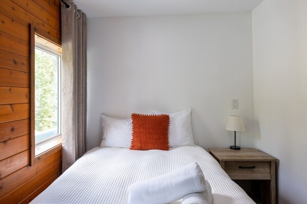 A bedroom with a wooden wall, a window, a bed with white bedding and an orange accent pillow, a bedside table with a lamp, and a nightstand with a drawer.