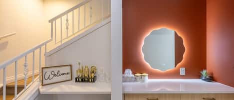 Una escalera con un cartel de 'Welcome', un espejo con un diseño de pared único y un tocador con encimera de madera.