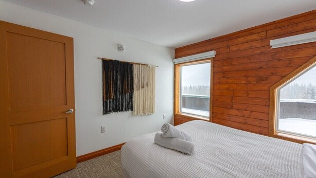 Une chambre avec un plafond en bois, une grande fenêtre, un lit recouvert d’une couette blanche et une porte.