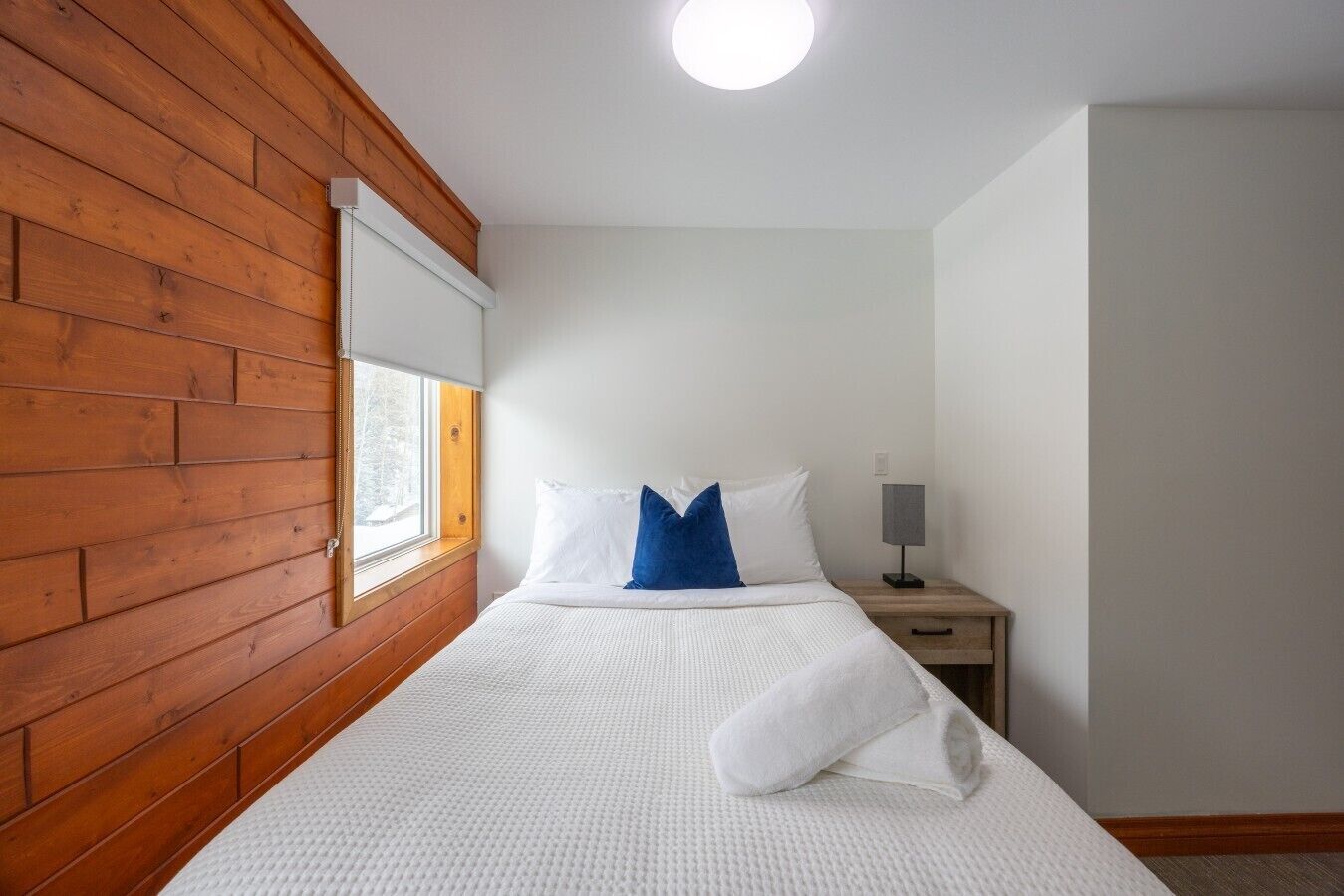 Habitación de hotel con pared de madera, cama con ropa blanca y almohada azul, mesita de noche con lámpara y ventana con vistas a árboles.