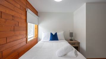 Habitación de hotel con pared de madera, cama con ropa blanca y almohada azul, mesita de noche con lámpara y ventana con vistas a árboles.