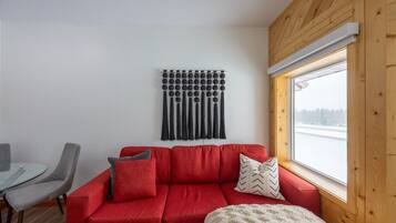 Un sofá rojo con cojines, una mesa de vidrio y sillas en una habitación con techo de madera y ventana con vista a un cuerpo de agua.