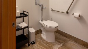 Une petite salle de bain privée équipée de toilettes, d’un sèche-mains, d’une poubelle et d’une étagère contenant des articles de toilette.