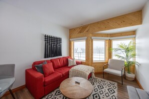 Een moderne woonkamer met een rode bank, een klein rond tafeltje en een witte fauteuil. Er is een groot raam met uitzicht op een besneeerd landschap.