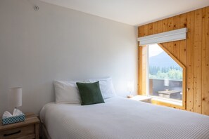 A bedroom with a bed, a nightstand, a lamp, a window with a view of greenery, and a wooden ceiling.