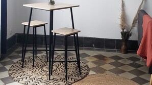 Ein Zimmer mit Schachbrettmuster-Boden, einem runden gewebten Teppich, einem Tisch mit zwei Stühlen und Wandregalen mit Dekorationen.