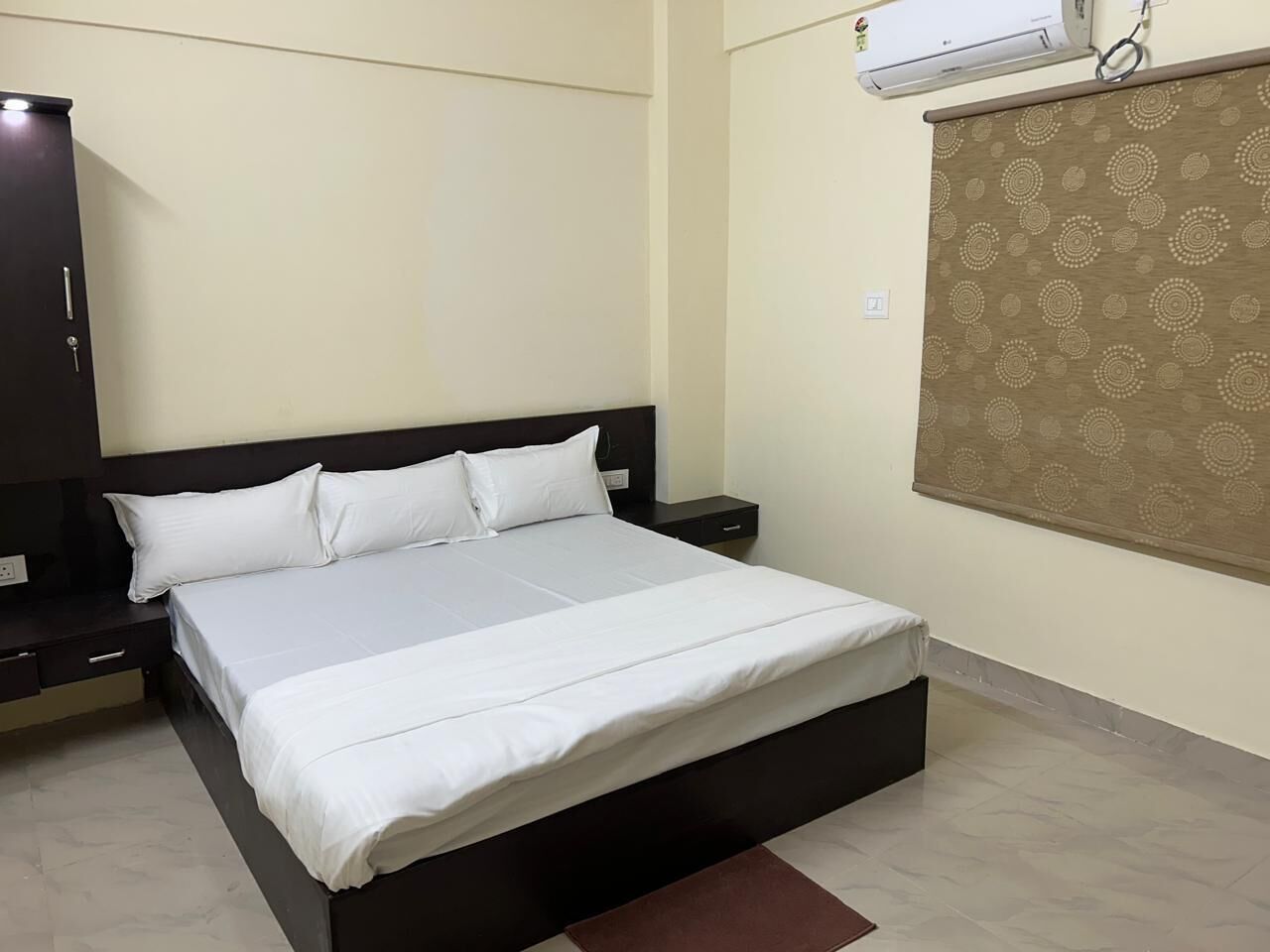 A bed with white bedding, a dark headboard, a nightstand with a lamp, and a wall-mounted air conditioner.