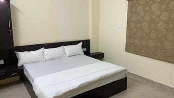 A bed with white bedding, a dark headboard, a nightstand with a lamp, and a wall-mounted air conditioner.