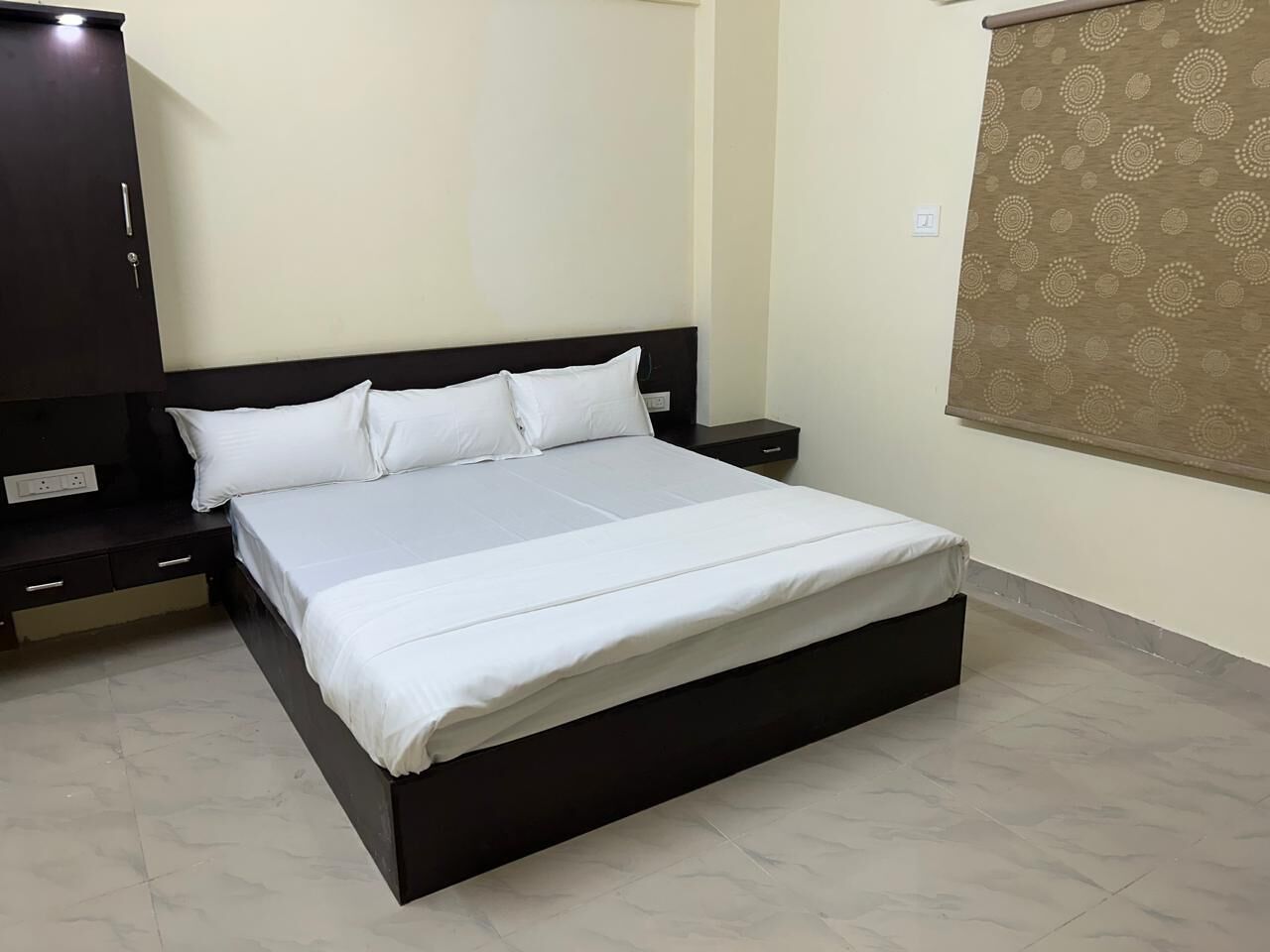 Uma cama de solteiro com cabeceira escura, mesa de cabeceira, guarda-roupa e cortina estampada.