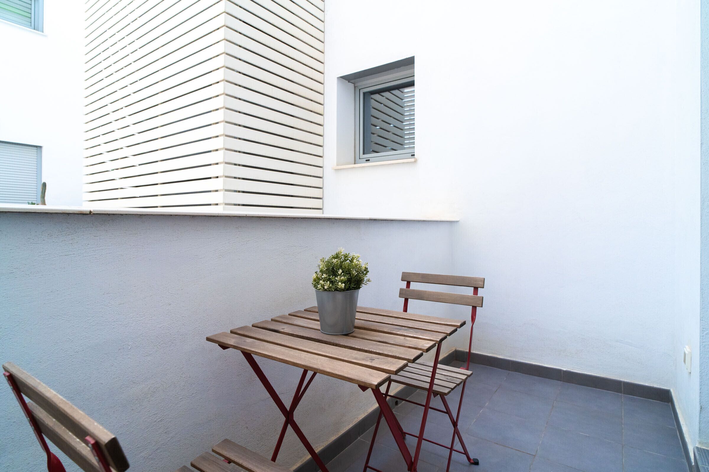 Une petite terrasse extérieure avec une table et des chaises en bois, une plante en pot et une fenêtre avec des stores.
