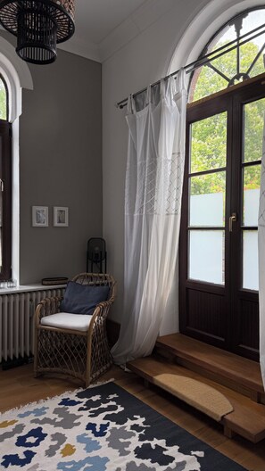 Ein Zimmer mit einem Korbstuhl, einer Bank, einem gemusterten Teppich, einem Heizkörper und einem Fenster mit Vorhang.