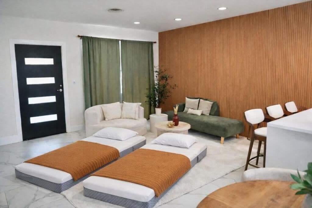 A modern hotel room with a sofa, two single beds, a green armchair, a small table, and a wooden accent wall.
