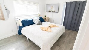 Un dormitorio con cama, ventana con persianas, televisor montado en la pared y cortina gris.