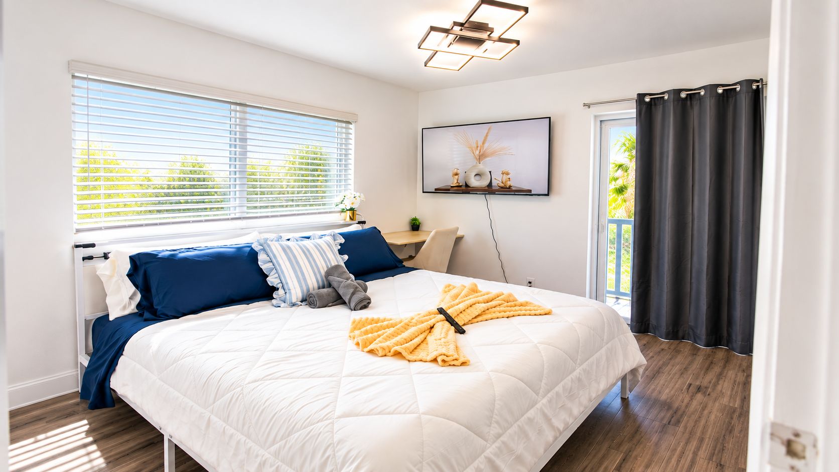 A bedroom with a bed, pillows, a bedside table, a window with blinds, a wall-mounted TV, and a door with curtains.