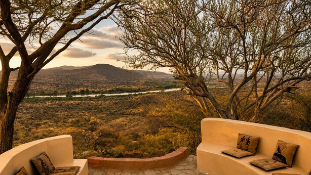 A curved outdoor seating area with stone flooring and cushions, overlooking a landscape with sparse vegetation and distant hills. - Nasuulu lodge (Isiolo)