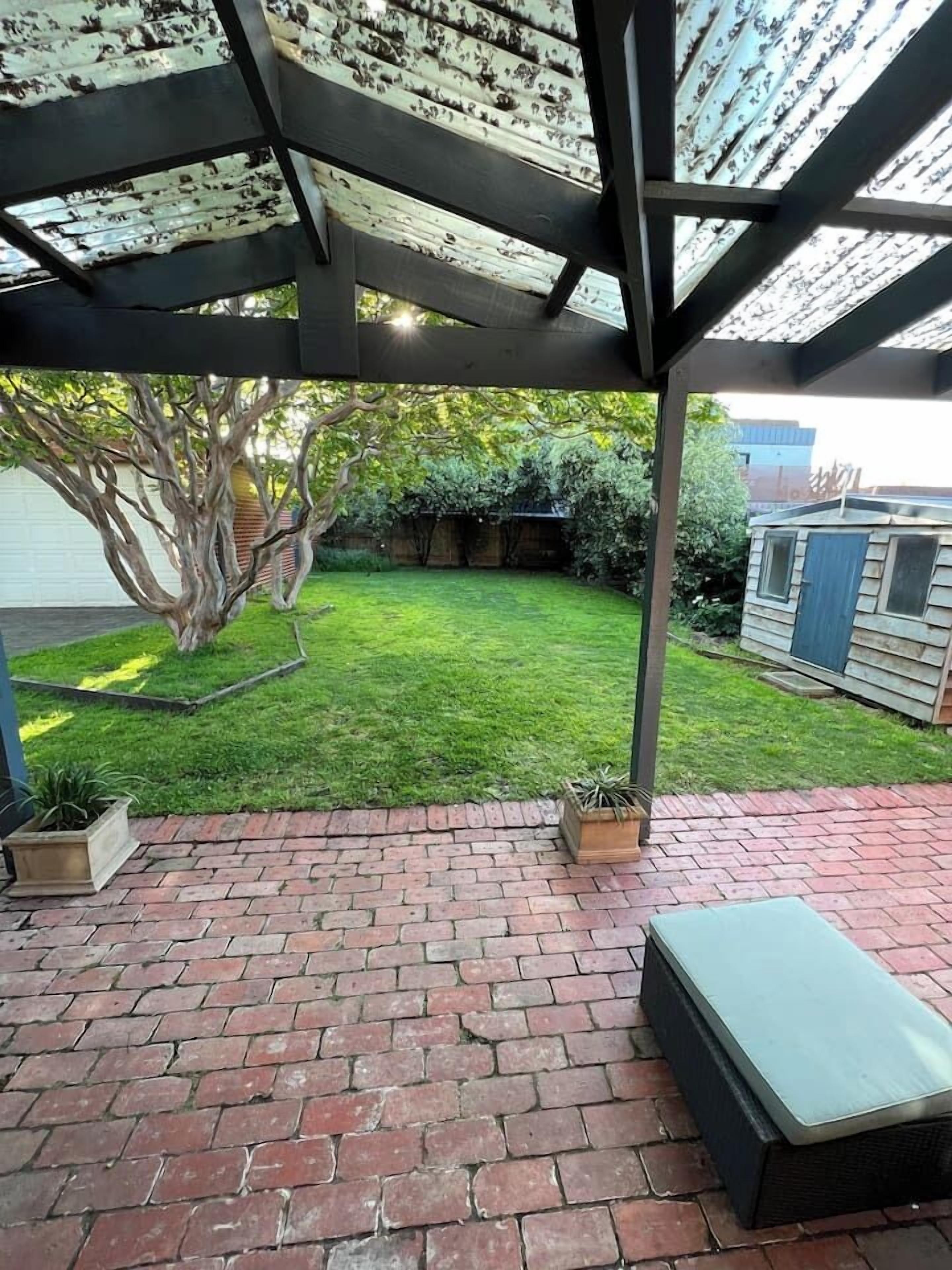 A covered patio with brick flooring, a bench, and a tree in the background.
