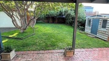A covered patio with brick flooring, a bench, and a tree in the background.