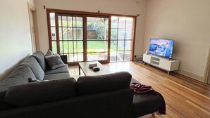 A living room with a grey sofa, a coffee table, a TV on a white stand, and a sliding glass door leading to a garden.