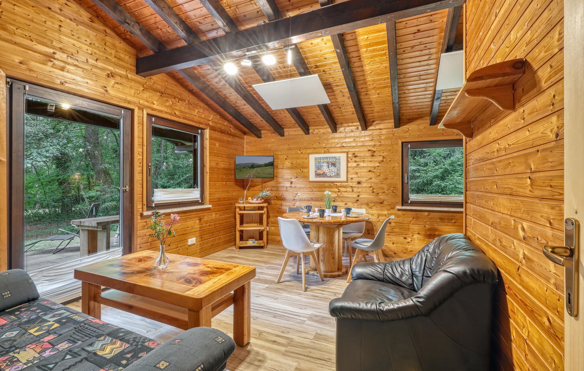 A wooden cabin interior with a dining area, a sofa, and a TV mounted on the wall.