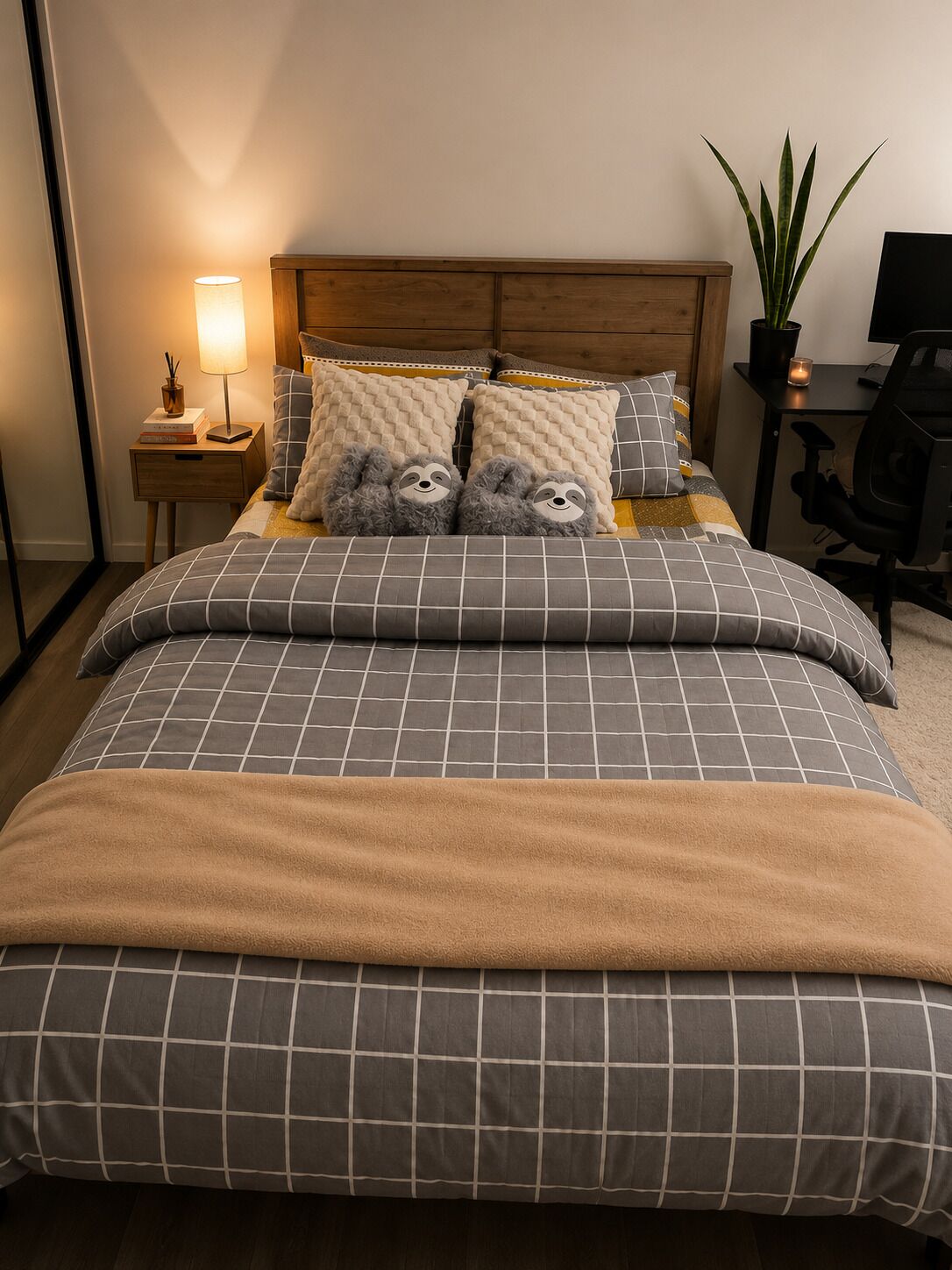 A neatly made bed with a checkered duvet and two pillows, a bedside lamp, a small wooden nightstand, a potted plant, and a desk with a chair.