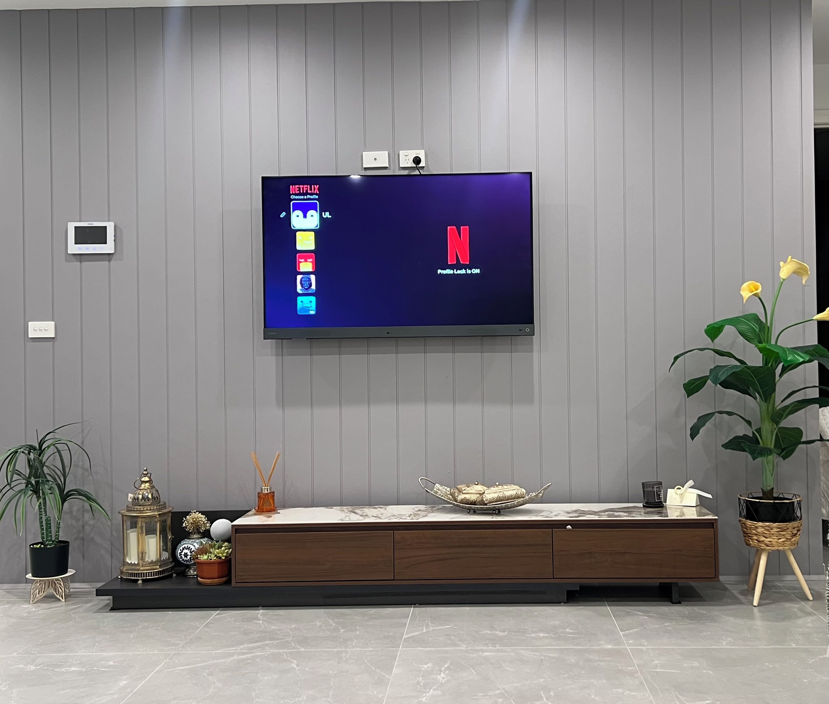 A modern living room with a flat-screen TV mounted on a grey wall, a wooden entertainment unit below, and a potted plant to the right.