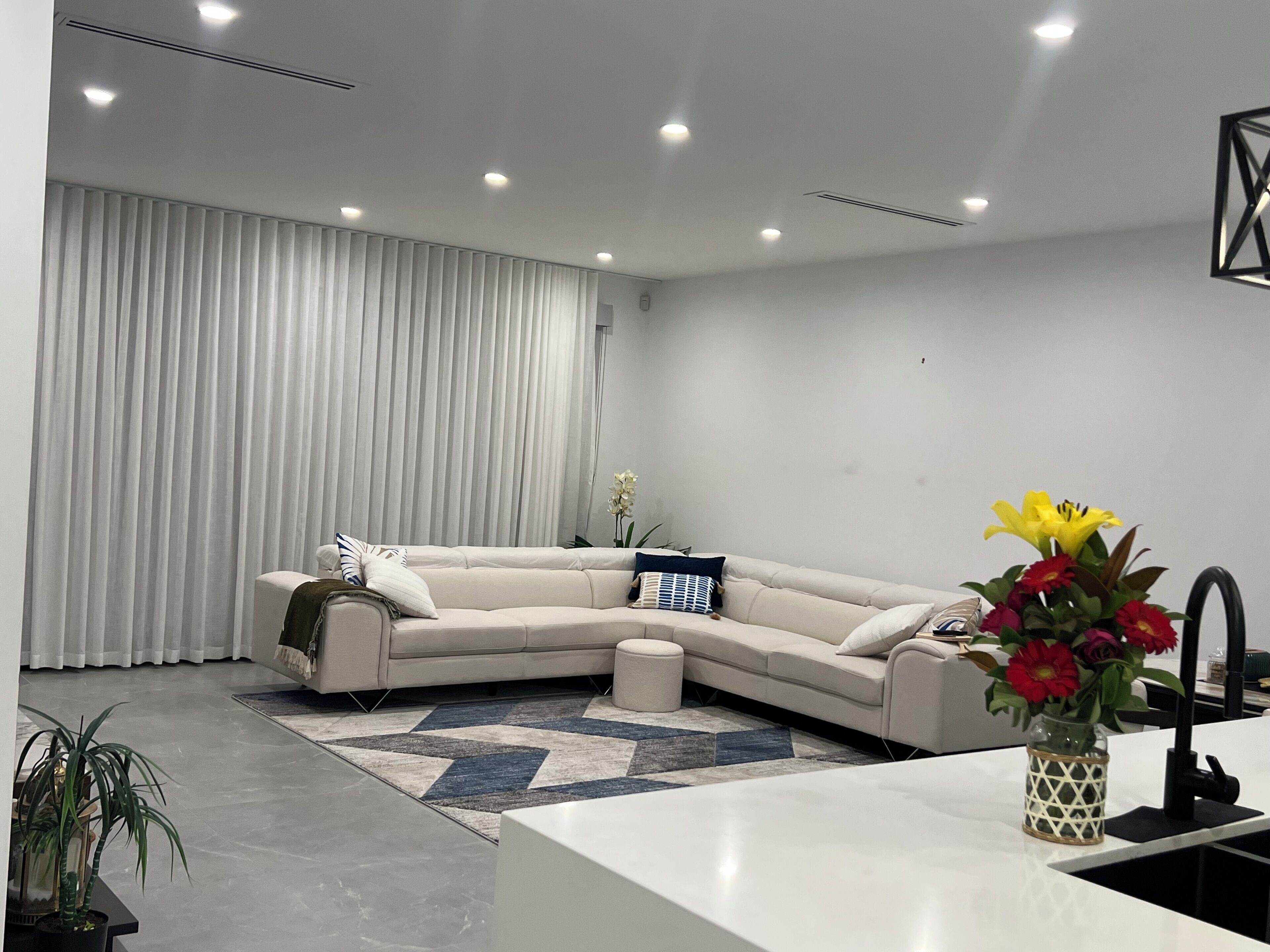 A modern living room with a sectional sofa, a small round coffee table, a kitchen area with a sink, and a potted plant.