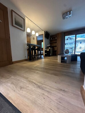 Een ruime woonkamer met houten vloer, een keukenhoek met eethoek en een groot raam met uitzicht op een gebouw.