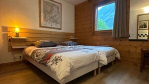 A bedroom with a wooden bed, a bedside table with a lamp, a window with curtains, and a framed picture on the wall. - Chamonix Town Centre Apartment with Free Parking (Chamonix-Mont-Blanc)