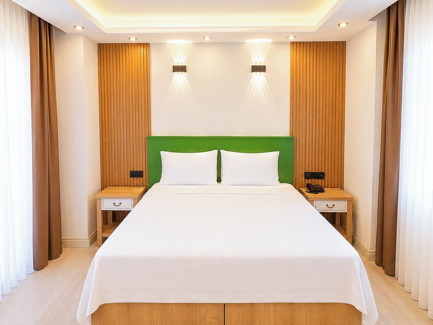 A hotel room with a large bed, two bedside tables, a green headboard, and wall-mounted lighting.