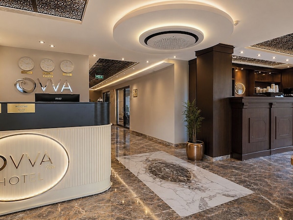 Uma área de recepção de hotel com design moderno, destacando um balcão de recepção, relógios exibindo a hora em Londres, Istambul e Tóquio, e um balcão com o nome do hotel.