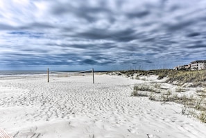 Een strand met een volleybalnet, zandduinen en een gebouw op de achtergrond.