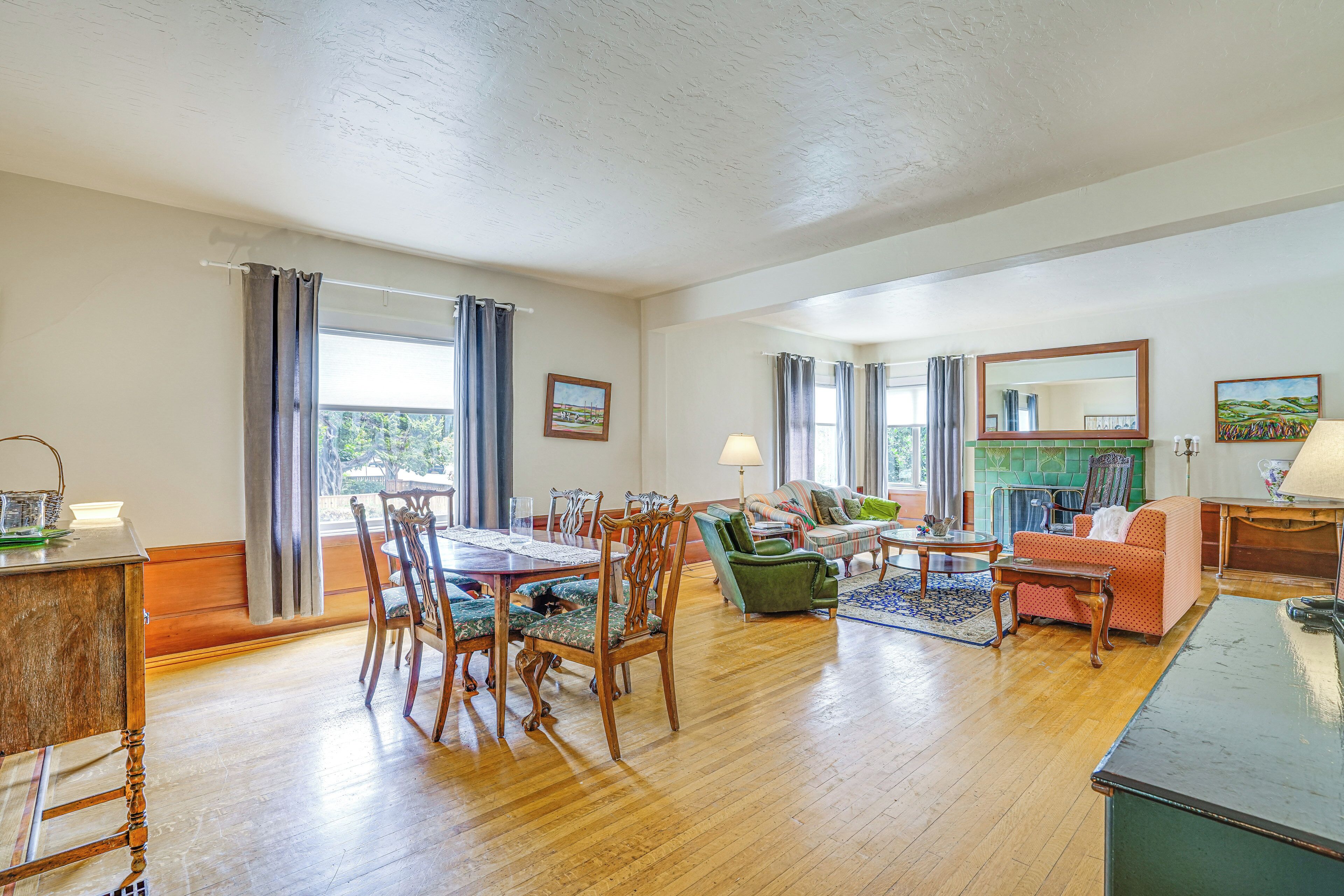 A spacious living room with a dining area, wooden flooring, and large windows with curtains.