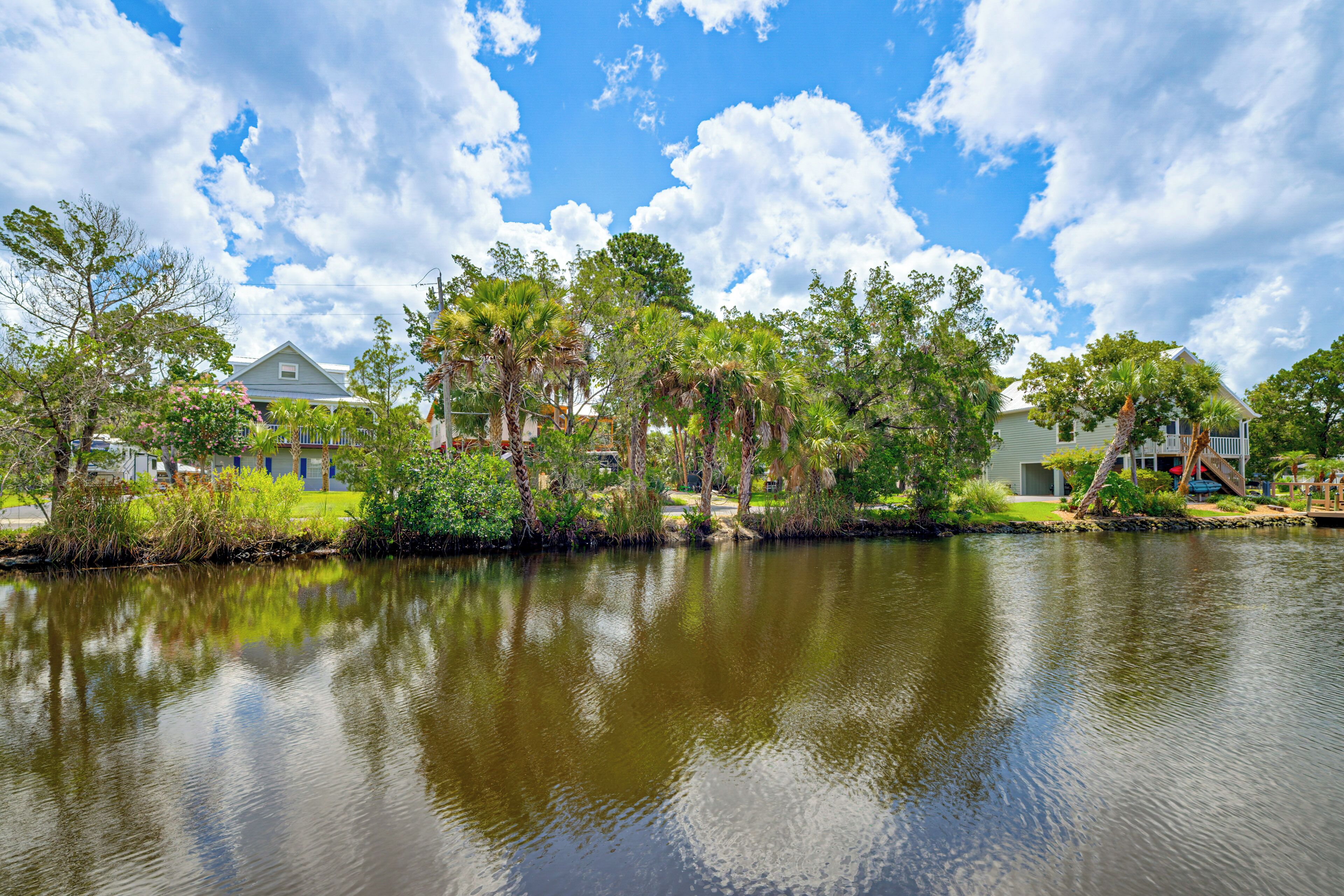 A calm lake with reflections of trees and houses, surrounded by greenery and clear skies.