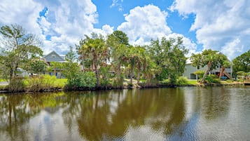 A calm lake with reflections of trees and houses, surrounded by greenery and clear skies.