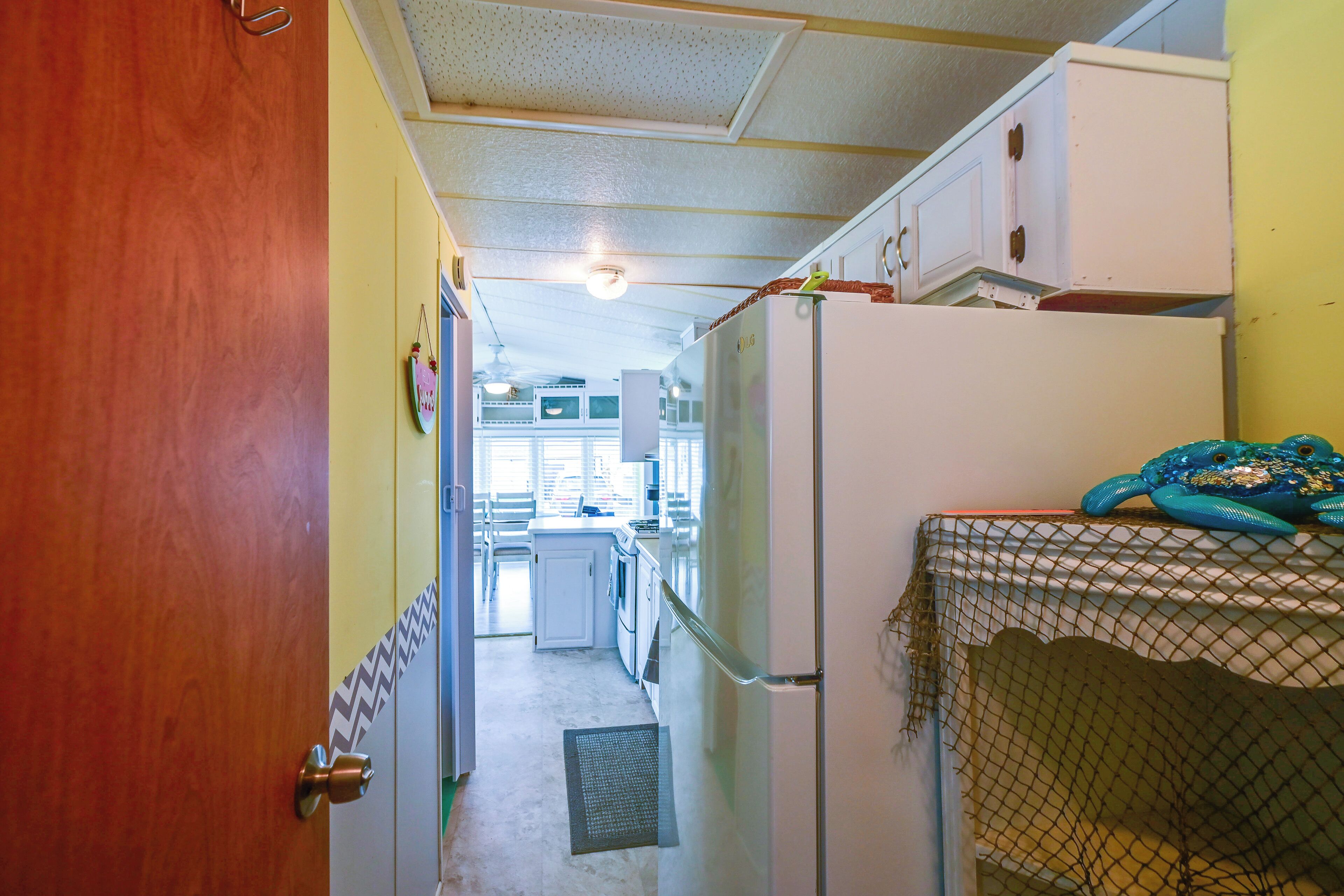 A narrow hallway with a wooden door on the left, a white refrigerator on the right, and a kitchen area in the background.