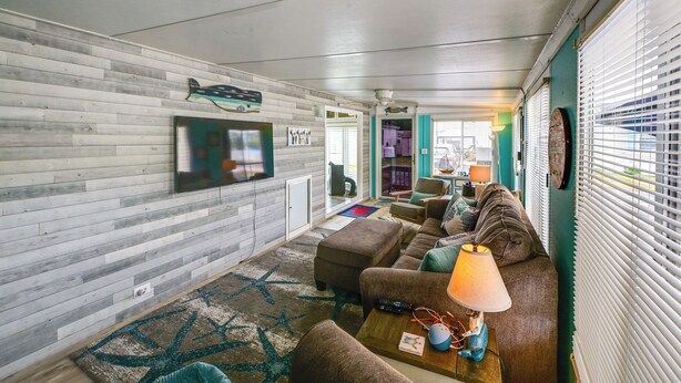 Ein Wohnzimmer mit Sofa, Hocker und einer Lampe. Ein Fernseher ist an der Wand montiert und es gibt ein Fenster mit Jalousien.