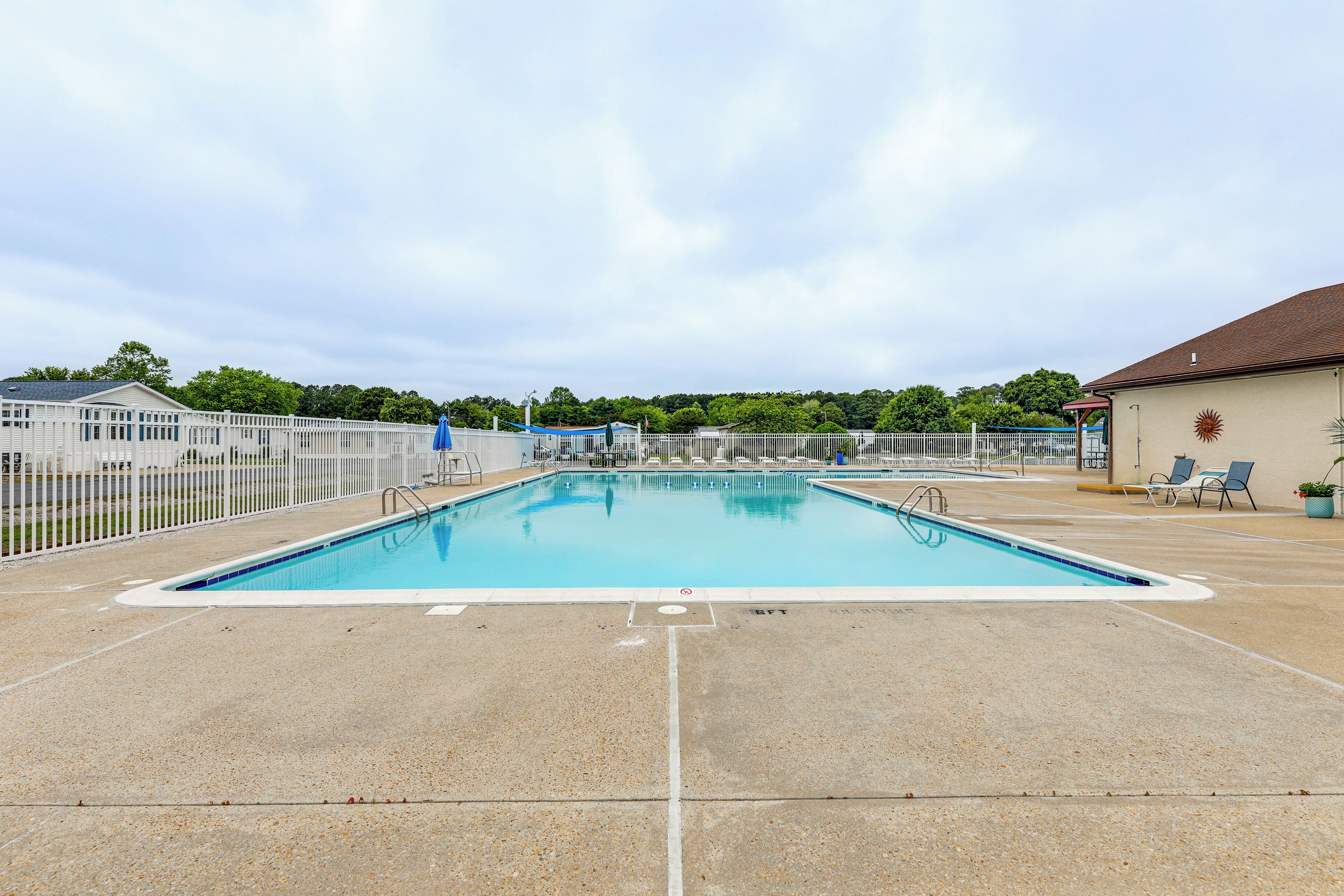 Una piscina rectangular con un borde blanco, rodeada por una cerca blanca, y un edificio con techo marrón al fondo.