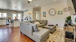 A modern living room with a sectional sofa, a ceiling fan, a kitchen area with white cabinets, and a decorative wall clock. - Pool, Games, Fenced Yard! Extended Stay in Buckeye (Buckeye)