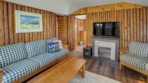 Ein Holzblockhaus-Interieur mit einem karierten Sofa, einem Kamin, einem Fernseher und einem gerahmten Landschaftsbild.