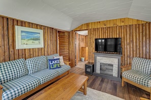 Ein Holzblockhaus-Interieur mit einem karierten Sofa, einem Kamin, einem Fernseher und einem gerahmten Landschaftsbild.