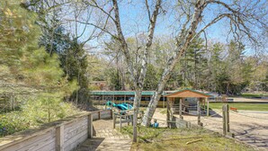 Eine Holzterrasse führt zu einem Pavillon mit Dach, umgeben von Bäumen und einem Gebäude im Hintergrund.