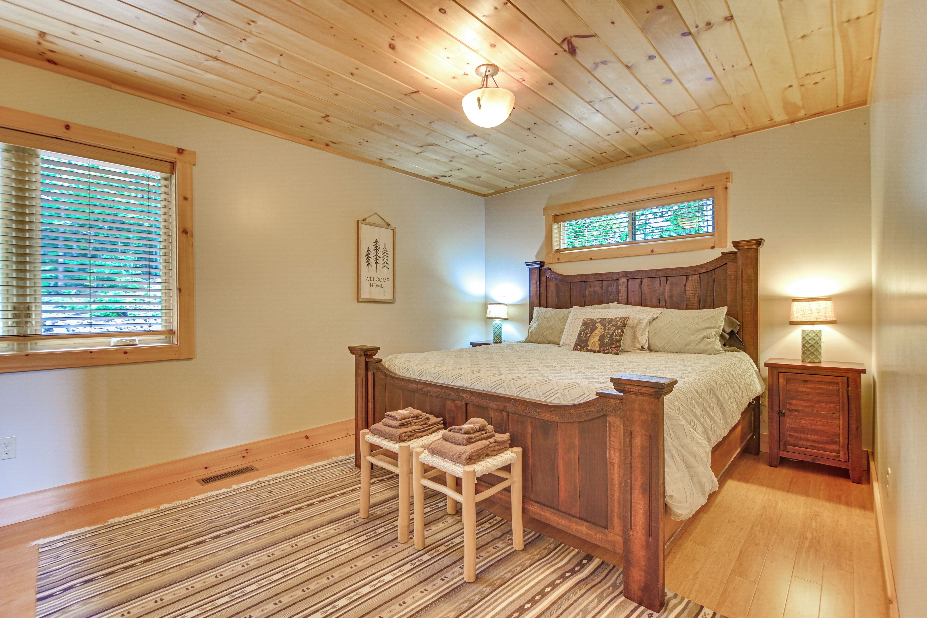 A bedroom with a wooden bed, two nightstands, a window with blinds, a wall-mounted light fixture, and a rug.
