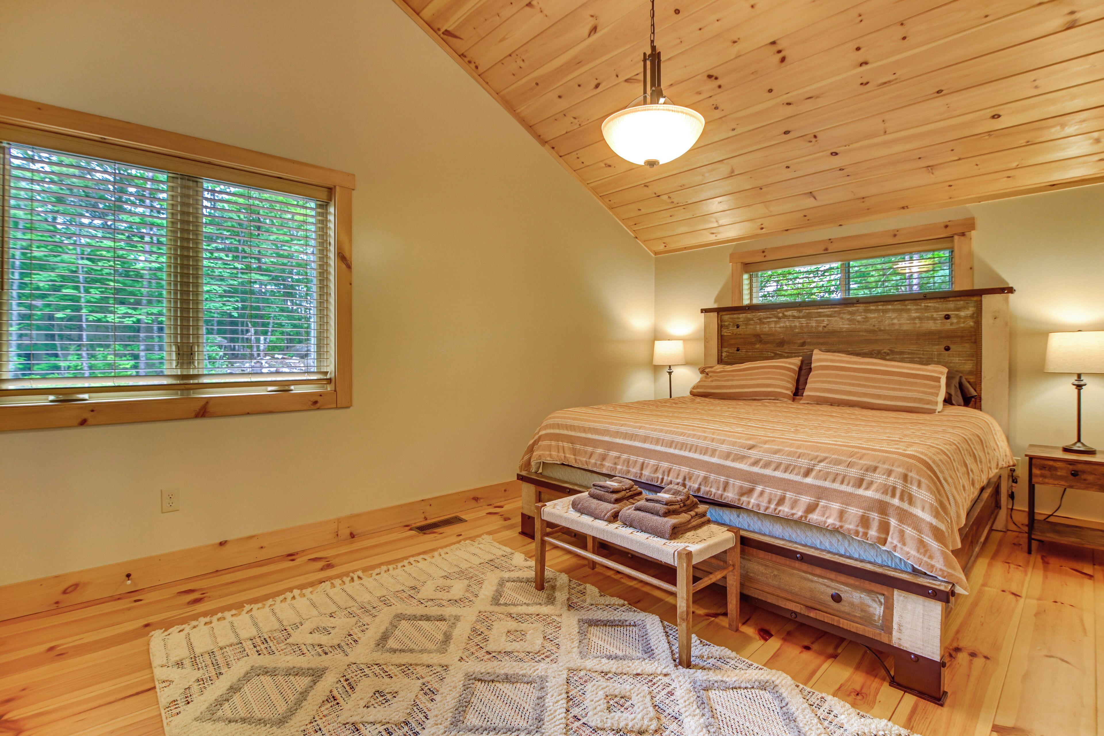 A bedroom with a wooden ceiling, a bed with a striped bedspread, a bench with towels, a patterned rug, and a window with blinds.