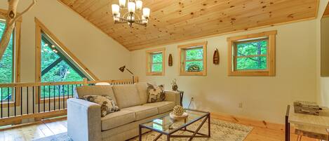 A spacious living room with a wooden ceiling, a glass coffee table, a sofa, and a view of the outdoors through large windows.