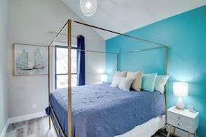 Ein Bett mit Baldachin, ausgestattet mit einer blauen Tagesdecke, ein Nachttisch mit Lampe und ein Gemälde von Segelbooten an der Wand.