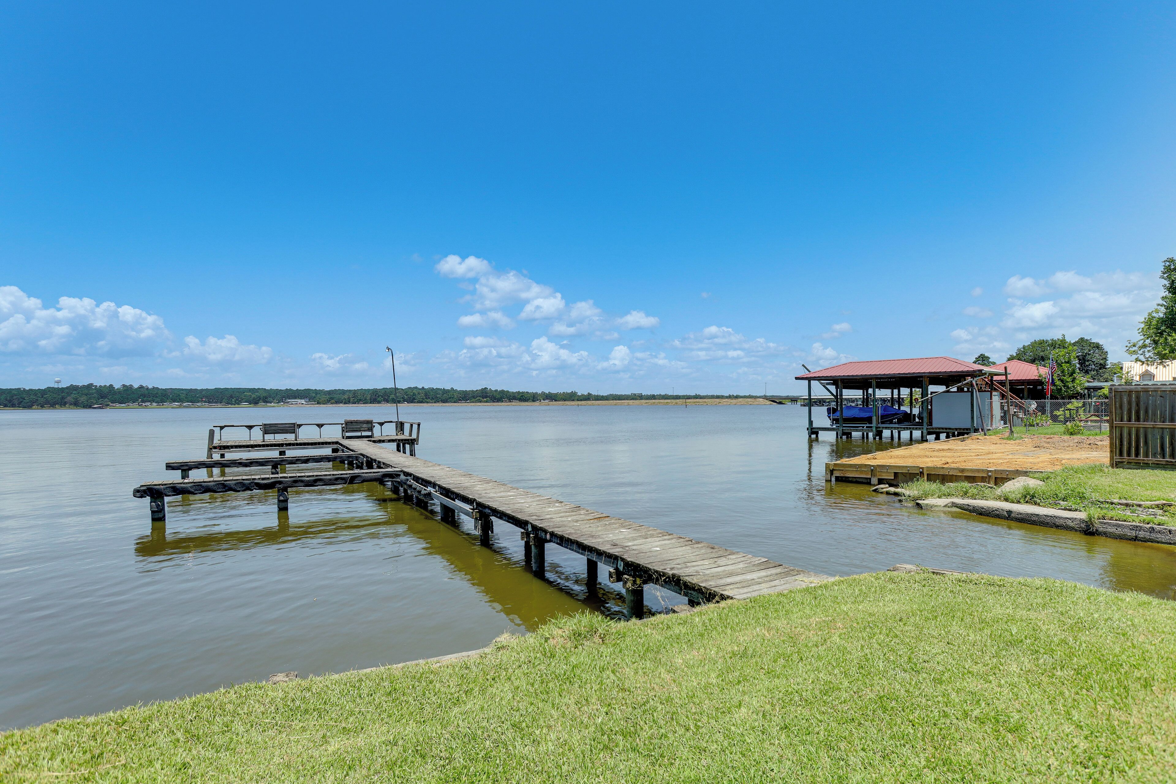 A lakeside dock with a wooden pier extending into the water, a small wooden structure with a red roof, and a grassy area in the foreground.