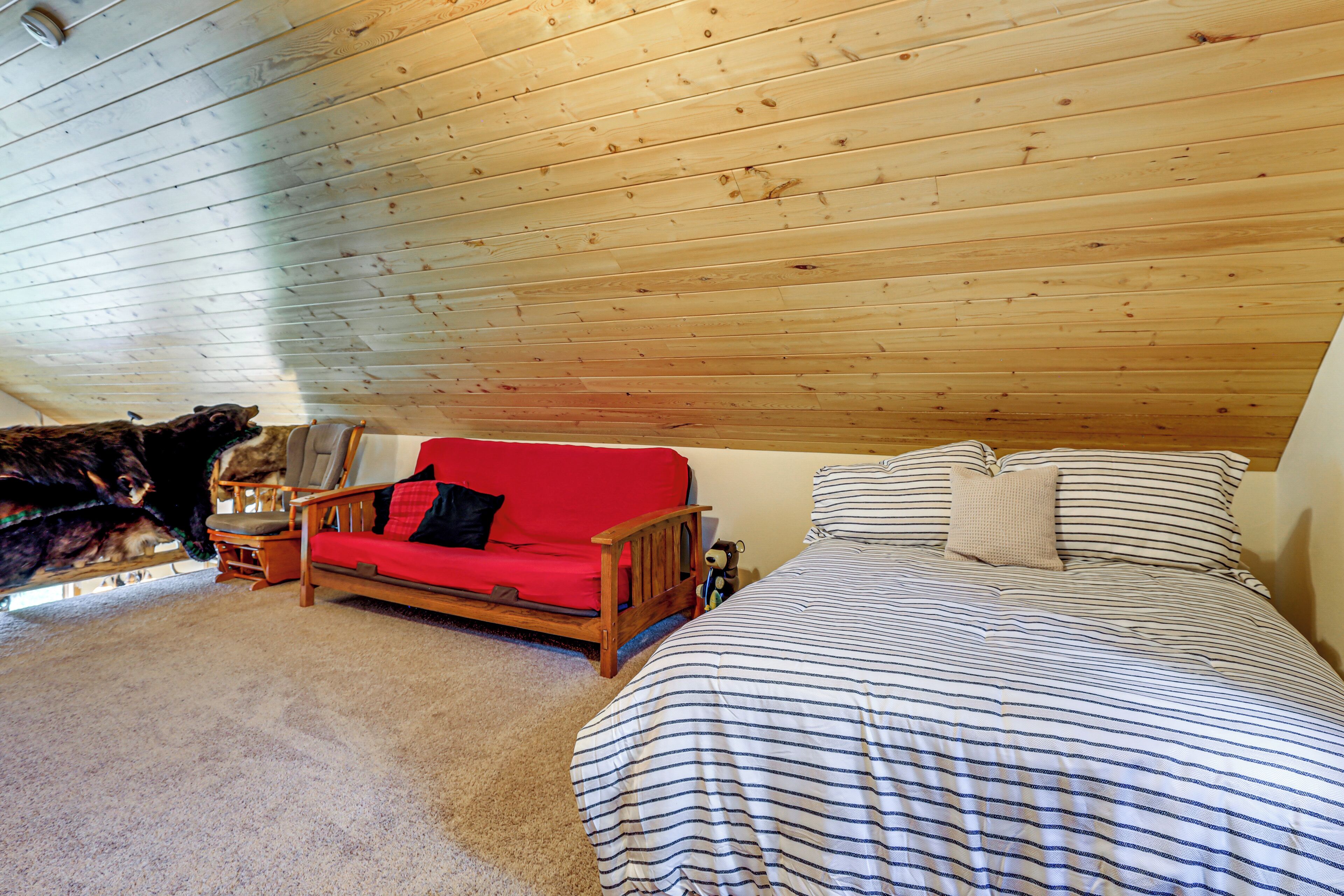 Quarto aconchegante com teto de madeira, sofá vermelho, cama listrada e um bicho de pelúcia no chão.