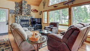 A cozy living room with a stone fireplace, comfortable seating, and large windows offering a view of the forest.