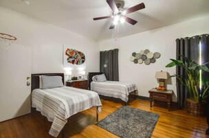 Habitación de hotel con dos camas, ventilador de techo, aro de baloncesto, una obra de arte en la pared, un adorno mural, una mesita de noche, una lámpara y una planta en maceta.